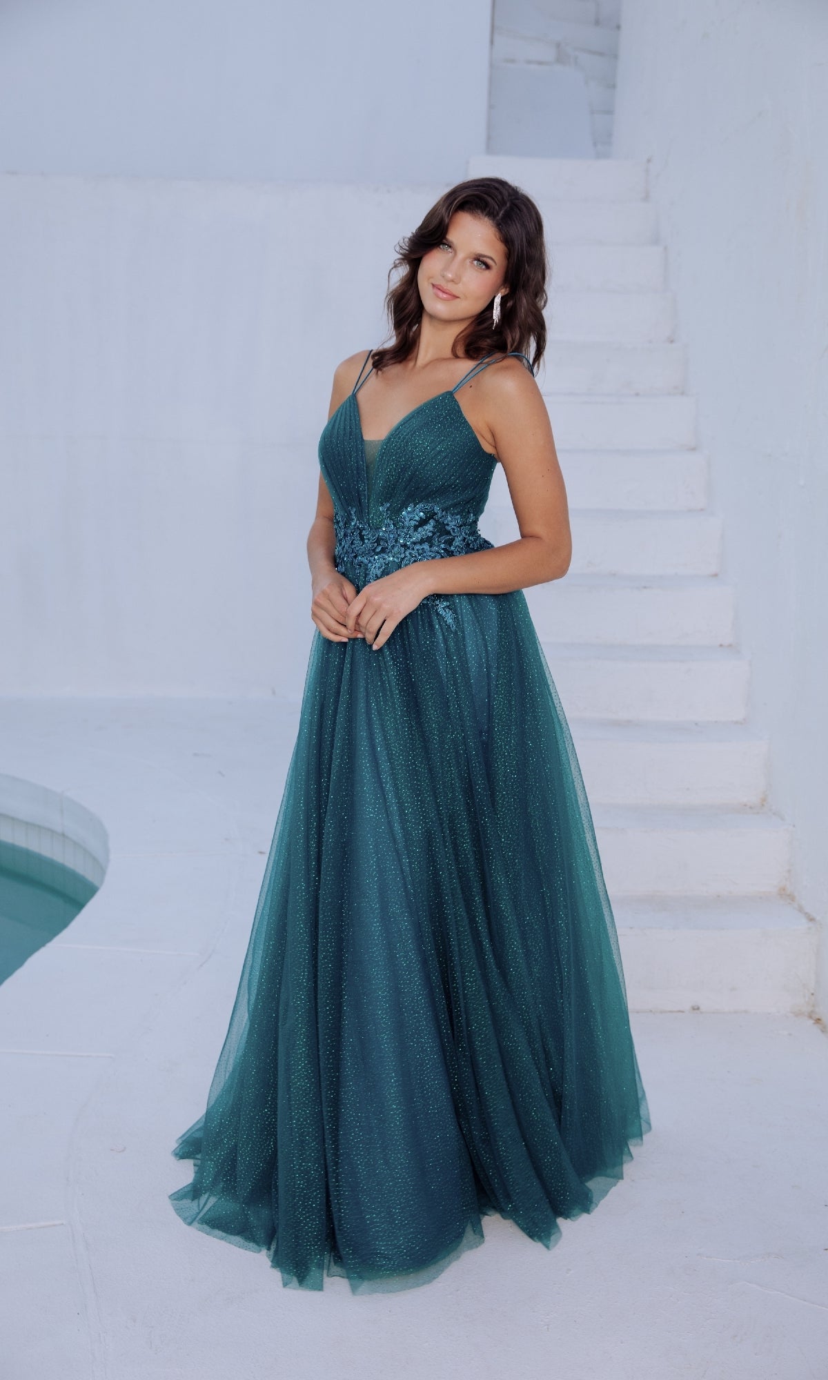 A woman in the Eureka 9797 Lace-Up Long Shimmer Prom Dress stands near white stairs and a curved pool.